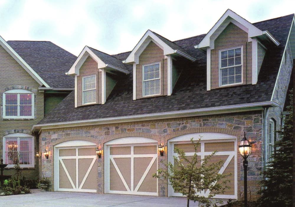 Three tan carriage-style garage doors on a stone home with dormer windows